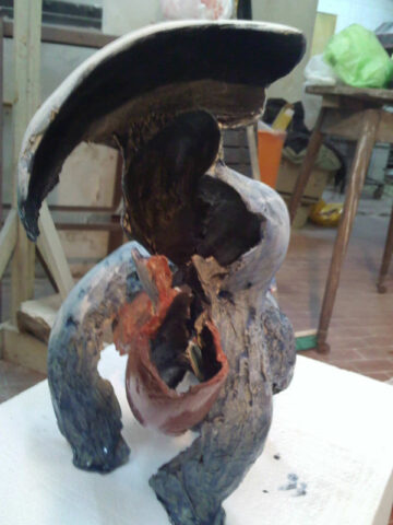 Escultura abstracta gris azulada con una forma de ala negra en la parte superior y una abertura que deja ver el interior rojo anaranjado, sobre un pedestal blanco.