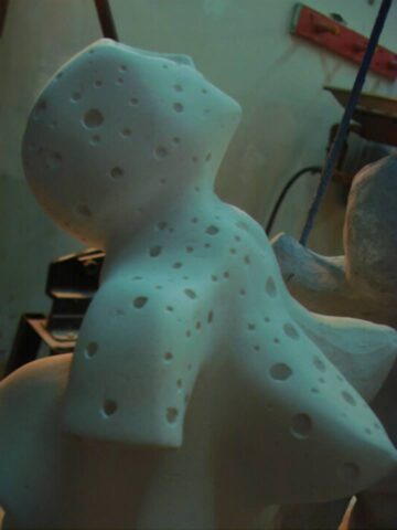 Escultura en yeso blanco con múltiples perforaciones circulares, forma humana estilizada vista de espaldas con los brazos extendidos, en un taller.
