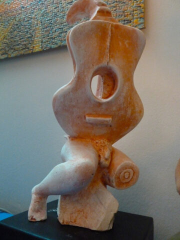 Escultura de cerámica color terracota con forma de guitarra estilizada, un orificio circular al centro y una espiral en la parte superior, sobre un pedestal negro.